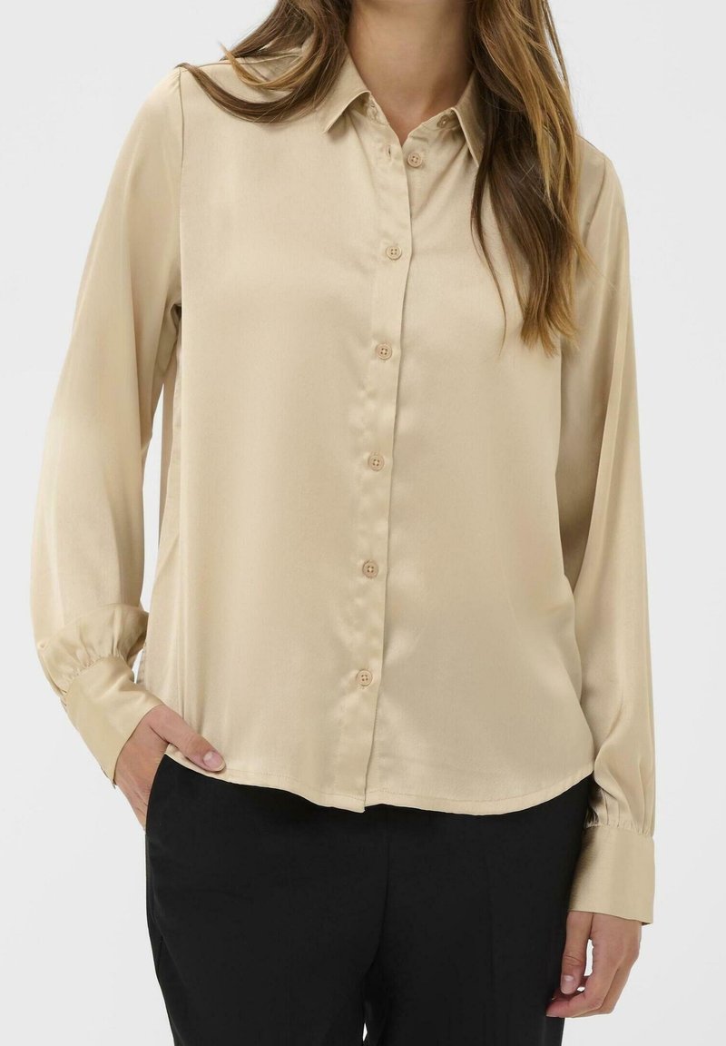 Femme portant une chemise beige à manches longues boutonnée avec col et un pantalon noir, une main dans la poche devant un fond uni.