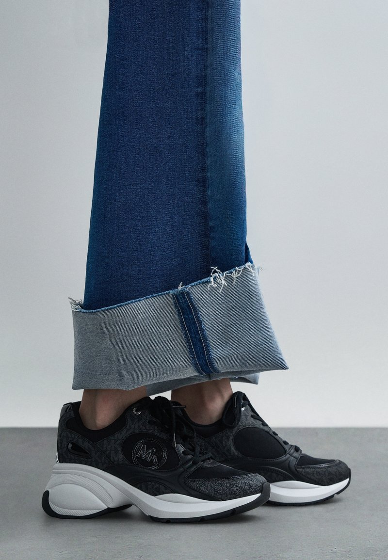 Black sneakers with textured design, grey and white accents. Accompanied by blue jeans with a frayed hem and lighter denim cuff.