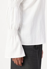 Top bianco a maniche lunghe con dettagli arricciati ai polsini, caratterizzato da una vestibilità rilassata e una texture liscia, abbinato a pantaloni in tessuto a spina di pesce scuri.