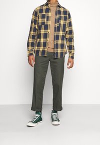 Camisa de cuadros en azul marino y amarillo, de manga larga, con dos bolsillos en el pecho; combinada con un suéter de cuello alto beige, pantalones verde oliva y zapatillas verdes.