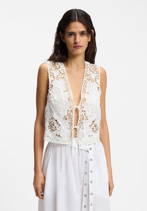 Γυναίκα που φοράει λευκό αμάνικο δαντελένιο γιλέκο δεμένο μπροστά και λευκή φούστα με ψηλή μέση και κουμπιά, σε μονόχρωμο φόντο.