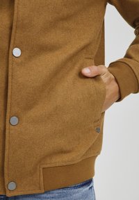 Chaqueta de mezcla de lana marrón con un acabado texturizado, botones a presión plateados, bolsillos laterales y puños y dobladillo acanalados. Presenta una etiqueta de logo sutil.