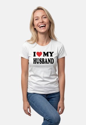 Hymyilevä vaaleatukkainen nainen, jolla on pitkät hiukset ja valkoinen T-paita, jossa lukee "Rakastan miestäni" punaisella sydämellä, yhdistettynä sinisiin farkkuihin.