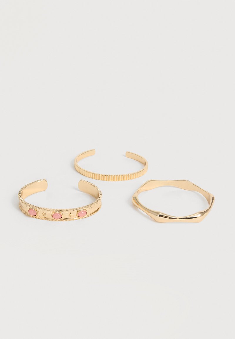Τρία ανοιχτά βραχιόλια σε χρυσό τόνο· το ένα με ροζ πέτρες και υφασμένα άκρα, το ένα με κάθετες ραβδώσεις και το ένα με ακανόνιστο εξάγωνο σχήμα.