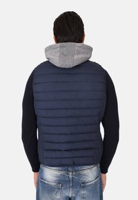 Gilet imbottito navy con texture a trapuntatura, cappuccio grigio e taglio aderente. Indossato sopra una maglietta a maniche lunghe scura, abbinato a jeans in denim chiaro.