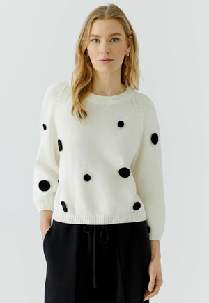 Blonde vrouw die een witte gebreide trui met zwarte cirkeldetails en een zwarte broek draagt, staand tegen een effen lichte achtergrond.