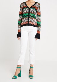 Kvinna som bär en flerfärgad zigzag-mönstrad kavaj, vita uppvikta byxor och gröna klacksandaler med ankelremmar.