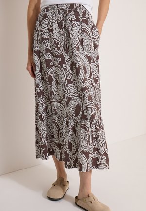 Person iført en brun og hvid paisleymønstret midinederdel, hvid top og beige slip-onsko, stående indendørs.