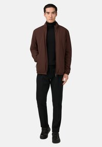 Braune leichte Jacke mit Stehkragen, schwarzer Rollkragenpullover, schwarze Hose und schwarze Sneaker. Glatte Textur, taillierter Schnitt.