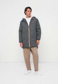 Manteau gris matelassé avec fermeture éclair jaune, capuche et doublure en fausse fourrure. Associé à un pantalon beige et des baskets blanches, porté par un mannequin masculin.