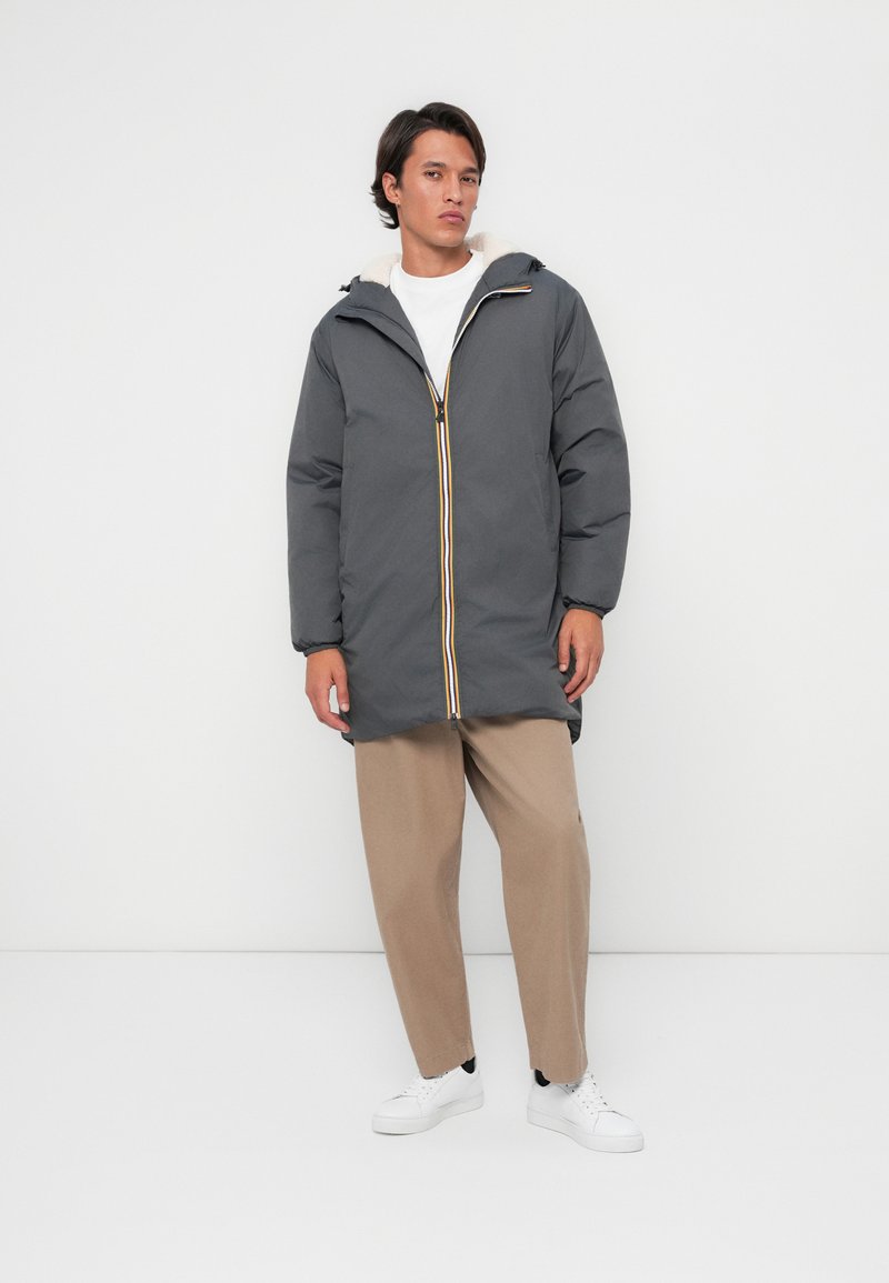 Polstret grå frakke med gul lynlås, hætte og faux pelsforing. Paireret med beige bukser og hvide sneakers, båret af en mandlig model.