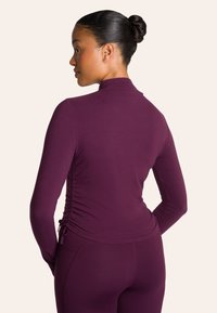 Langarmtop in diep bordeauxrood met hoge hals, ruching aan de zijkanten en een soepele textuur. Gecombineerd met bijpassende getailleerde leggings.