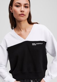Felpa bianca e nera con scollatura a V, caratterizzata da un design a spacco distintivo, logo ricamato "KARL LAGERFELD JEANS" e vestibilità rilassata.