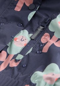 Material rezistent la apă, cu un fundal închis și modele colorate de copaci și fețe zâmbitoare. Include un buzunar cu fermoar și detalii cu nasturi.