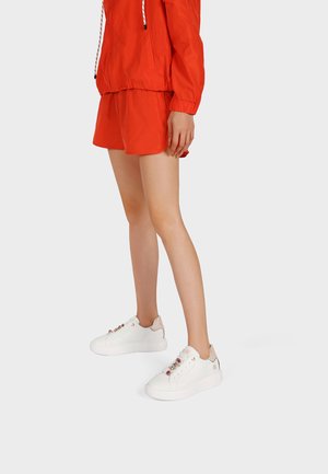 Gambe con indosso scarpe da ginnastica bianche con dettagli gioiello e una giacca a vento rossa coordinata con pantaloncini su uno sfondo chiaro.