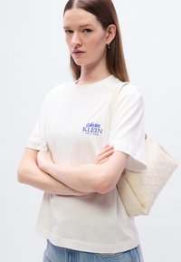 Άσπρο βαμβακερό T-shirt με μπλε λογότυπο "calvin KLEIN NEW YORK", συνδυασμένο με υφασμάτινη μπεζ τσάντα ώμου και ανοιχτό μπλε τζιν παντελόνι.