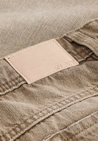 Bež denim tkanina savijena s vidljivim smešnim kožnim etiketom urezanim s "NEX." Ima teksturiranu površinu i šivene ivice.