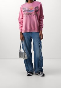 Pink sweatshirt with graphic design depicting palm trees and sunset; paired with light blue baggy cargo jeans and a silver handbag.