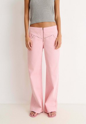 Femme portant un pantalon large taille haute rose clair avec fermeture éclair devant et petits clous, associé à un débardeur court gris et des sandales roses.
