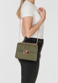 Bolso de mano verde oliva con un diseño estructurado, detalles de hardware en dorado y una correa de cadena. Material similar al cuero con acentos texturizados.