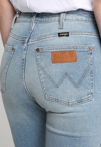 Jeans de mezclilla azul claro con cintura alta, bolsillos traseros con una etiqueta de cuero marrón y detalles de costura visibles.