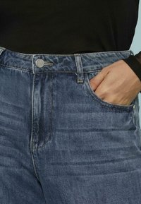 Jeans in denim blu chiaro con vita alta, tasche frontali e chiusura a bottone argentato. Il tessuto presenta leggere scoloriture e texture.