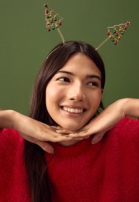 Gouden hoofdband met feestelijk boomontwerp, met rode en groene kerstballen en een ster, tegen een groene achtergrond. Model draagt een rode, pluizige trui.