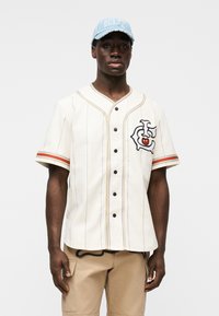 Λευκό πουκάμισο στυλ μπέιζμπολ με μαύρα κουμπιά και λεπτές ρίγες, με πολύχρωμο γραφικό σχέδιο στο στήθος. Ανοιχτό μπλε καπέλο φορεμένο στο κεφάλι.