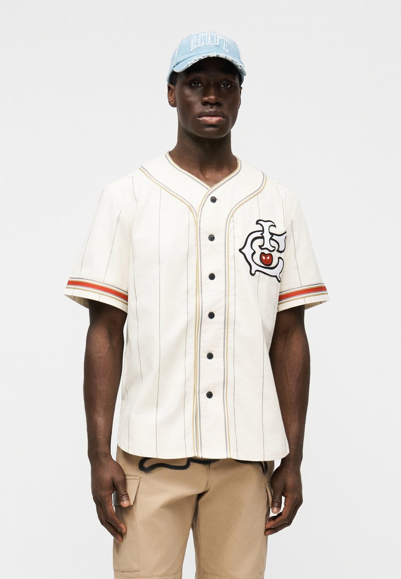 Λευκό πουκάμισο στυλ μπέιζμπολ με μαύρα κουμπιά και λεπτές ρίγες, με πολύχρωμο γραφικό σχέδιο στο στήθος. Ανοιχτό μπλε καπέλο φορεμένο στο κεφάλι.