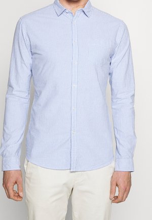 Homme portant une chemise à manches longues à rayures verticales bleu clair et blanc avec un pantalon blanc, debout devant un fond uni.