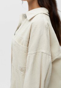 Beige ribjacket med krave, der har vertikal ribbet tekstur, forlods lomme og sølvknap på kraven.