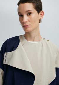 Beige en marineblauwe asymmetrische jas met een brede kraag, textuurstof en knoopaccenten nabij de schouder. Lichtgewicht ontwerp.