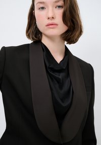 Musta räätälöity bleiseri, jossa on leveä ruskea satiininen shaalikaulus ja silkkinen musta drapeerattu toppi alla. Mallilla on sileä ihonväri.