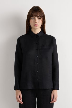 Vrouw met stijlbruin haar, gekleed in een zwart knopjeslangemouwen overhemd en bijpassende zwarte broek, staand tegen een effen witte muur.