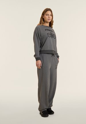 Giovane donna in piedi che indossa una felpa grigio scuro e pantaloni della tuta abbinati con scarpe da ginnastica nere su uno sfondo chiaro uniforme.