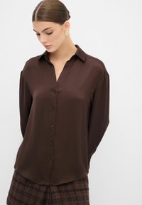 Blusa marrón de manga larga en una tela suave, con un cierre de botones en la parte delantera y un cuello clásico, combinada con pantalones de patrón a cuadros.
