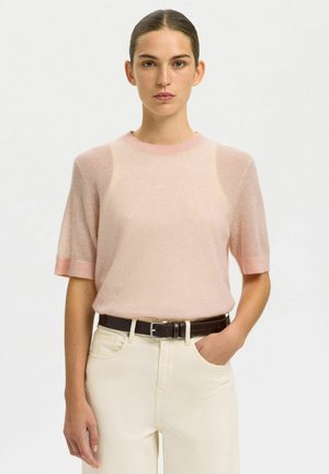 Femme aux cheveux foncés attachés porte un pull rose clair à manches courtes et un pantalon crème taille haute avec une ceinture foncée, une main dans la poche.
