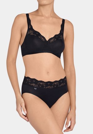 Femme portant un soutien-gorge noir orné de dentelle et un slip taille haute assorti, debout avec les mains détendues le long du corps sur un fond gris.