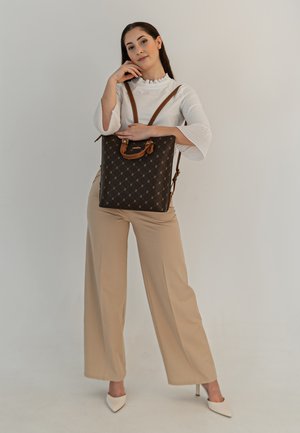 Bruine geblokte tote bag met een plat ontwerp, leren accenten en verstelbare banden, vastgehouden door een persoon in een witte top en beige broek.