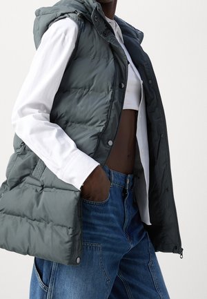 Person iført en mørkegrå dunvest over en hvid cropped skjorte og blå denimjeans med hænderne i lommerne.