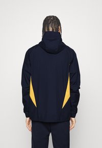 Persoon met gevlochten haar, gekleed in een marineblauwe jas met gele, driehoekige zijpanelen, staand met de rug naar de kijker tegen een effen achtergrond.