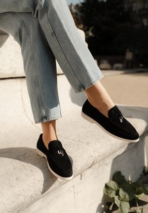 Schwarze Wildleder Slipper mit einer weißen Gummisohle, ausgestattet mit einem oberen Riemen und metallischen Logo-Akzenten. Getragen mit hellblauen cropped Jeans.