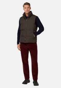 Moški temno rjav puhovni brezrokavnik z visokim ovratnikom, sprednjim zadrgo in stranskimi žepi, v kombinaciji z burgundskimi kabelskim hlačami in črnimi supergami.