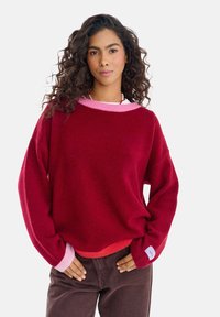 Czerwony sweter z różowymi akcentami, z okrągłym dekoltem, o luźnym kroju, z długimi rękawami, miękkiej teksturze, zaokrąglonym dołem, noszony na brązowych spodniach, z detalem w postaci metki.