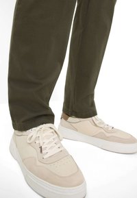 Beige sneakers med ruskind og læderdetaljer, med perforeret tå, tyk hvid sål og markante snørebånd. Olivengrønne bukser med en ren afslutning.
