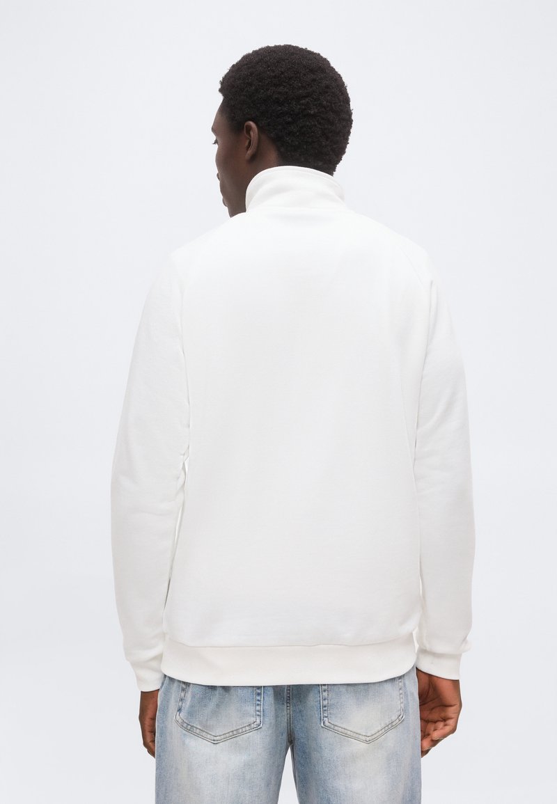 Homme aux cheveux bouclés portant un sweat-shirt blanc à col roulé et un jean bleu clair, vu de dos sur un fond uni.