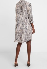 Kobieta w sukience w długości do kolan, z długim rękawem w wężowy wzór, z kołnierzykiem, stojąca tyłem do kamery, w brązowych butach na obcasie.