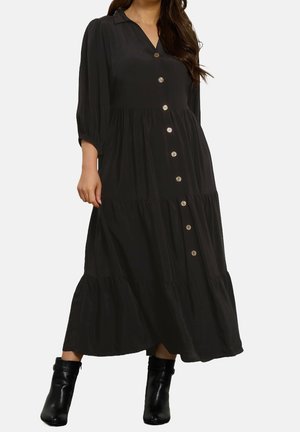 Femme portant une longue robe noire à boutons avec manches trois-quarts et des bottines noires, debout devant un fond uni.