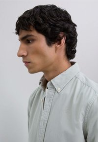Camisa verde claro de botones con cuello, textura sutil y un pequeño emblema en el lado izquierdo del pecho, llevada por una persona de perfil.