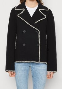 Zwarte dubbelzijdige blazer met contrasterende witte stiksels, gesneden revers en twee voorkantzakken; gemaakt van een gestructureerde stof.